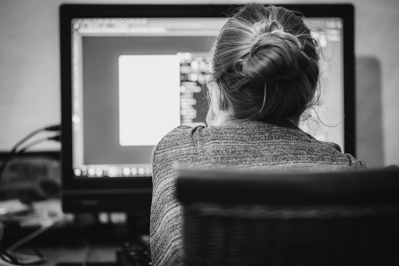 A person seen from behind sits at a desk working at a large computer monitor, in black and white